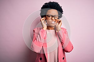 Young beautiful African American afro businesswoman with curly hair wearing pink jacket covering ears with fingers with annoyed