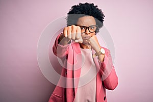 Young beautiful African American afro businesswoman with curly hair wearing pink jacket Punching fist to fight, aggressive and