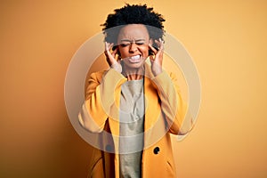 Young beautiful African American afro businesswoman with curly hair wearing yellow jacket covering ears with fingers with annoyed