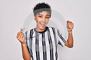 Young beautiful african american afro referee woman wearing striped uniform using whistle screaming proud and celebrating victory