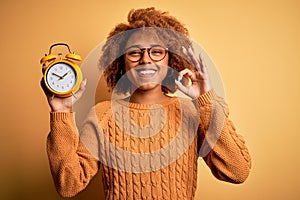 Young beautiful African American afro woman with curly hair holding vintage alarm clock doing ok sign with fingers, excellent