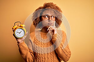 Young beautiful African American afro woman with curly hair holding vintage alarm clock serious face thinking about question, very