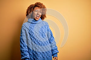 Young beautiful African American afro woman with curly hair wearing blue turtleneck sweater winking looking at the camera with