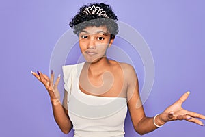 Young beautiful african american afro woman wearing tiara crown over purple background clueless and confused expression with arms
