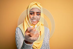 Young beautiful african american girl wearing muslim hijab over isolated yellow background Beckoning come here gesture with hand