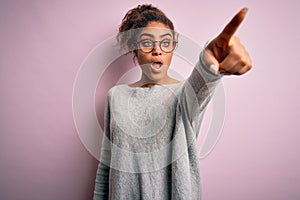 Young beautiful african american girl wearing sweater and glasses over pink background Pointing with finger surprised ahead, open