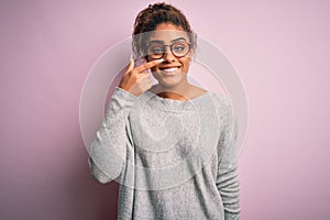 Young beautiful african american girl wearing sweater and glasses over pink background Pointing with hand finger to face and nose,