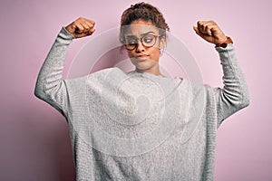 Young beautiful african american girl wearing sweater and glasses over pink background showing arms muscles smiling proud