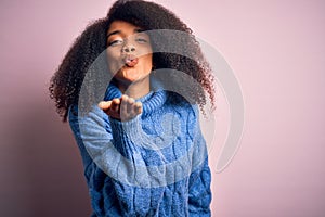 Young beautiful african american woman with afro hair wearing winter sweater over pink background looking at the camera blowing a