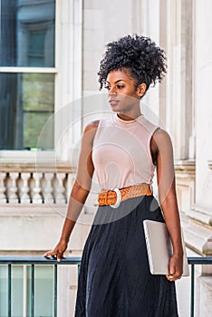 Young beautiful African American woman with afro hairstyle wearing sleeveless light color top, belt, black skirt, holding laptop