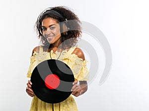 Young beautiful african american woman with headphones in yellow dress enjoying and dancing with vinyl record at white