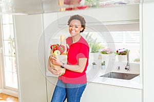 Young beautiful african american woman holding paper bag full of fresh healthy groceries at the kitchen