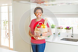Young beautiful african american woman holding paper bag full of fresh healthy groceries at the kitchen