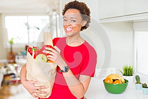 Young beautiful african american woman holding paper bag full of fresh healthy groceries at the kitchen