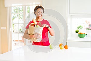 Young beautiful african american woman holding paper bag full of fresh healthy groceries leaving vegetables on the table