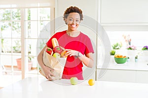Young beautiful african american woman holding paper bag full of fresh healthy groceries leaving vegetables on the table