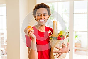 Young beautiful african american woman holding paper bag full of fresh healthy groceries and picking vegetables