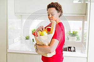 Young beautiful african american woman holding paper bag full of fresh healthy groceries and picking vegetables