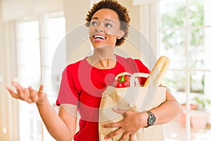 Young beautiful african american woman holding paper bag full of fresh healthy groceries and picking vegetables