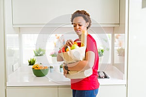 Young beautiful african american woman holding paper bag full of fresh healthy groceries and picking vegetables