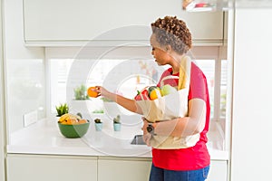 Young beautiful african american woman holding paper bag full of fresh healthy groceries and picking vegetables