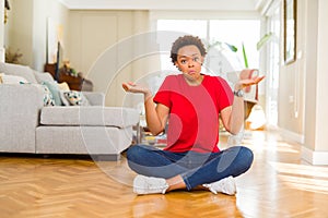 Young beautiful african american woman sitting on the floor at home clueless and confused expression with arms and hands raised