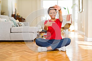 Young beautiful african american woman sitting on the floor at home smiling making frame with hands and fingers with happy face