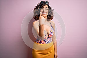 Young beautiful arab woman on vacation wearing swimsuit and sunglasses over pink background Beckoning come here gesture with hand