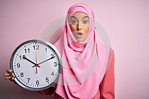 Young beautiful arab woman wearing pink muslim hijab doing countdown holding big clock scared in shock with a surprise face,