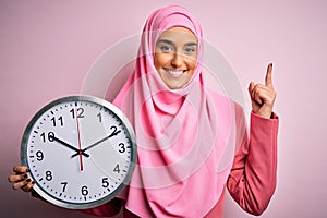 Young beautiful arab woman wearing pink muslim hijab doing countdown holding big clock surprised with an idea or question pointing