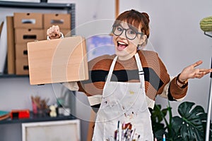 Young beautiful artist woman holding wooden case celebrating achievement with happy smile and winner expression with raised hand