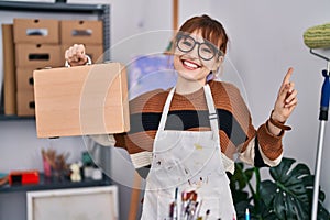 Young beautiful artist woman holding wooden case smiling happy pointing with hand and finger to the side