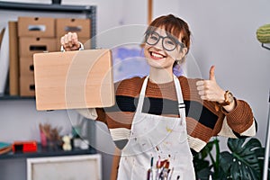 Young beautiful artist woman holding wooden case smiling happy and positive, thumb up doing excellent and approval sign