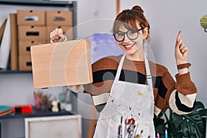 Young beautiful artist woman holding wooden case smiling with an idea or question pointing finger with happy face, number one