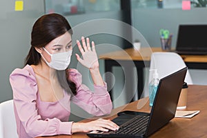 Young beautiful asian business woman wearing protective face mask greeting colleagues through video conferencing working in office