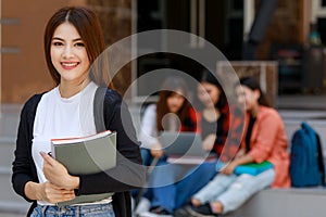 Young and beautiful Asian college student girls holding books, pose to camera with group of friends blur in background against