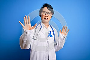 Young beautiful asian doctor girl wearing stethoscope and coat with blue cancer ribbon afraid and terrified with fear expression