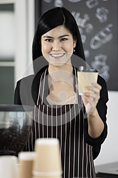 Young and beautiful Asian female barista wearing an apron standing in front of coffee machine. She holding offering paper coffee