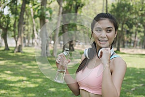 young beautiful asian fitness athlete woman holding drinking water after work out exercising at summer green park.