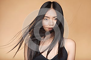 A young beautiful asian girl dressed in a black dress with natural makeup and shiny hair in the wind stands on a beige