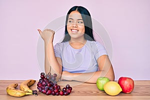 Young beautiful asian girl eating fresh and healthy fruit pointing thumb up to the side smiling happy with open mouth