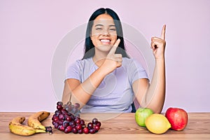 Young beautiful asian girl eating fresh and healthy fruit smiling and looking at the camera pointing with two hands and fingers to