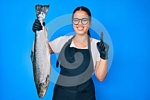 Young beautiful asian girl fishmonger selling fresh raw salmon smiling with an idea or question pointing finger with happy face,