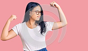 Young beautiful asian girl wearing casual clothes and glasses showing arms muscles smiling proud