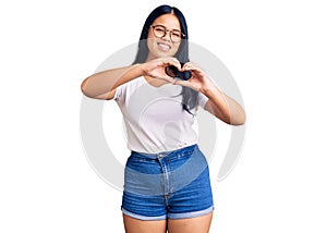 Young beautiful asian girl wearing casual clothes and glasses smiling in love doing heart symbol shape with hands