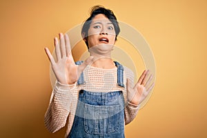 Young beautiful asian girl wearing casual denim overalls over isolated yellow background afraid and terrified with fear expression