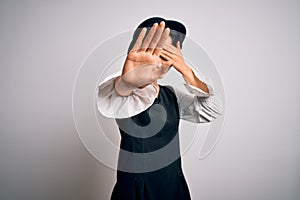 Young beautiful asian girl wearing casual dress and hat standing over isolated white background covering eyes with hands and doing
