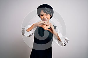Young beautiful asian girl wearing casual dress and hat standing over isolated white background smiling in love doing heart symbol