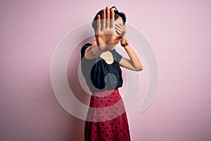Young beautiful asian girl wearing casual dress standing over isolated pink background covering eyes with hands and doing stop