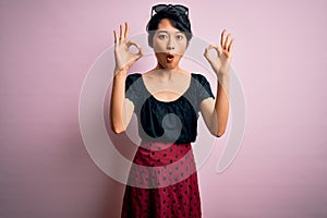 Young beautiful asian girl wearing casual dress standing over isolated pink background looking surprised and shocked doing ok